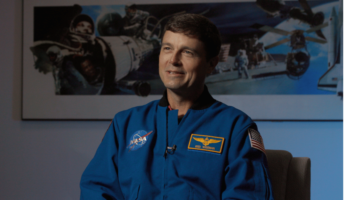 Astronaut in a blue NASA flight suit sits for an interview, smiling slightly. A name patch reading “Reid Wiseman” and a U.S. flag are visible on the suit. Behind him, a wall display shows images of spaceflight, including astronauts, spacecraft, and a space shuttle.