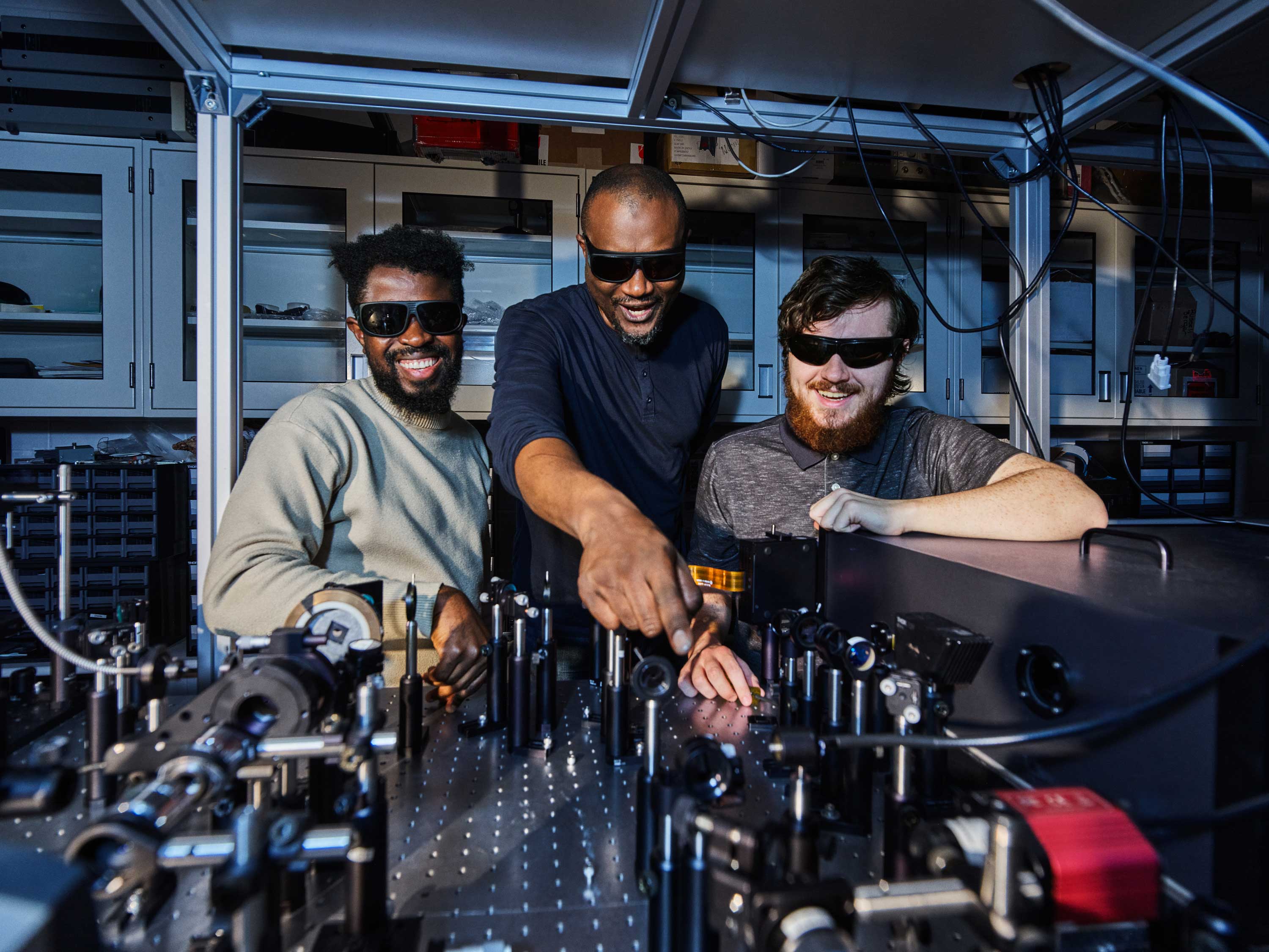 Three individuals wearing safety glasses are engaging with scientific equipment on a laboratory table. They appear to be working with precision instruments, possibly related to optics or physics. The setting is a modern lab with shelves and various apparatus in the background.