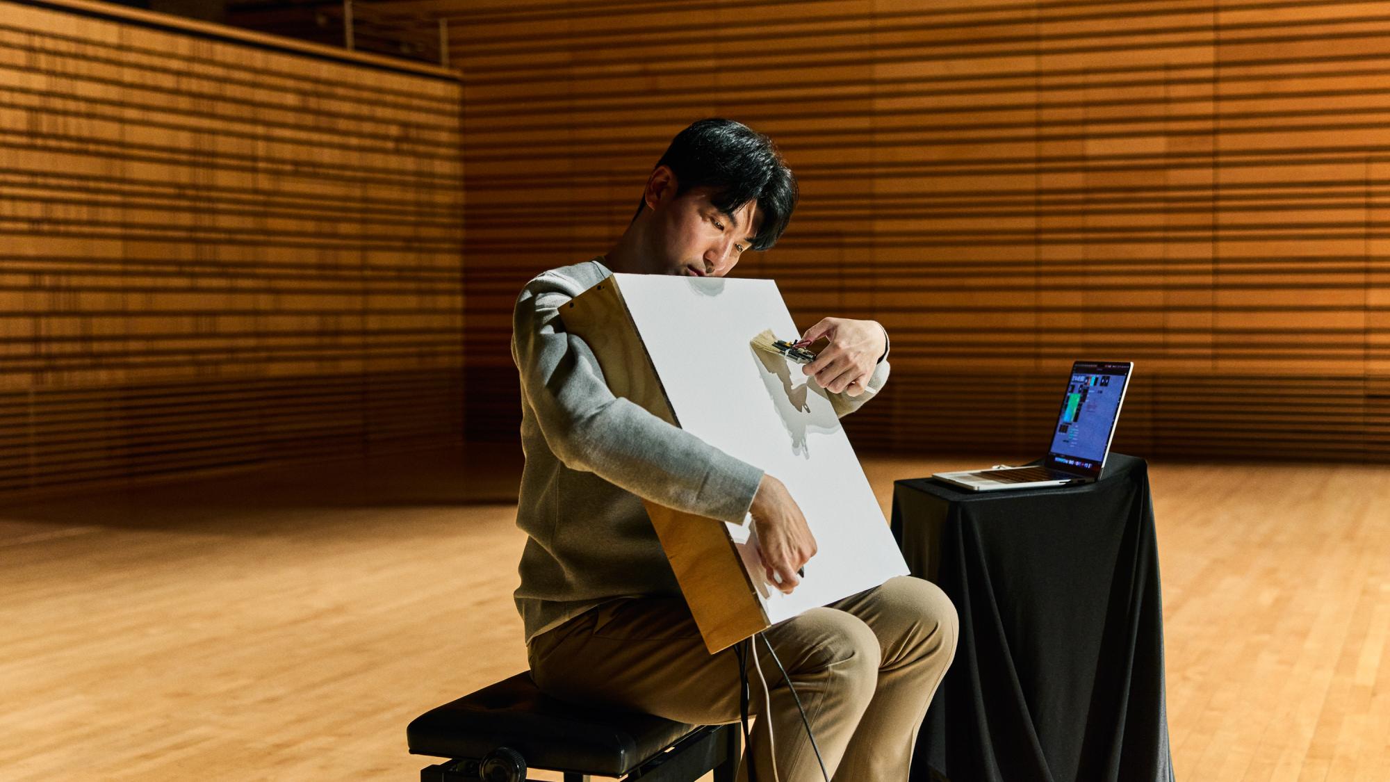 An individual sits on a bench on a large wooden stage, interacting with a white, rectangular electronic acoustic instrument. A laptop displaying software sits on a small table nearby, with ribbed wooden concert hall walls in the background.