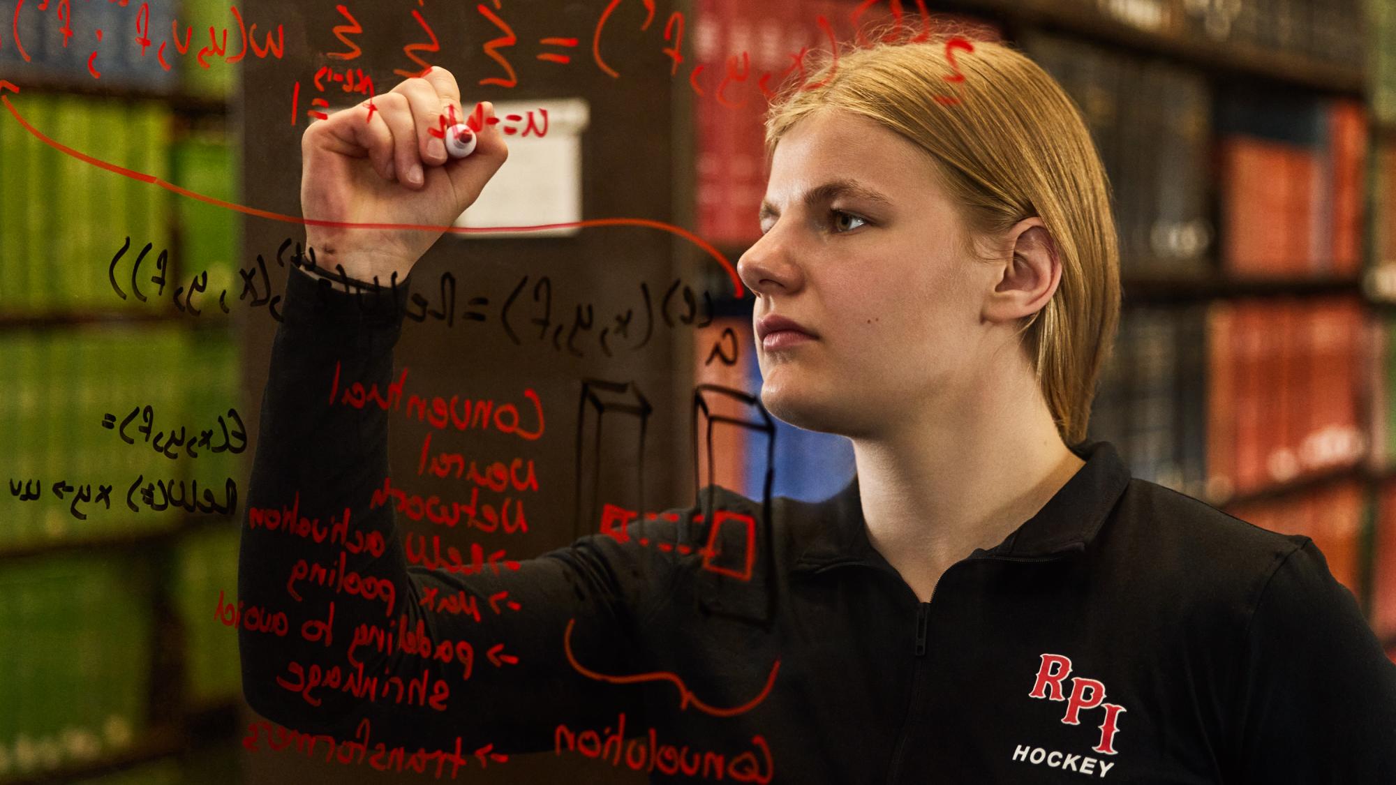 A student wearing an RPI Hockey pullover writes complex mathematical formulas and neural network diagrams in red marker on a clear glass board, with library bookshelves blurred in the background.