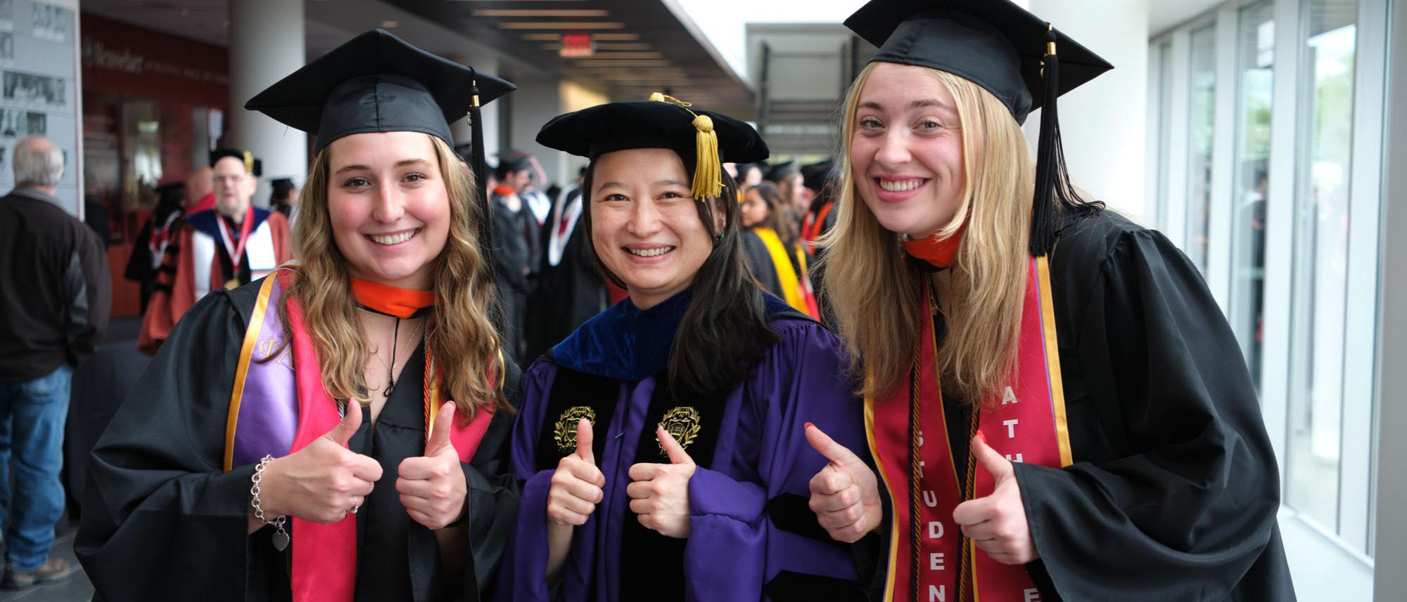 Three graduates in caps and gown pose with thumbs up
