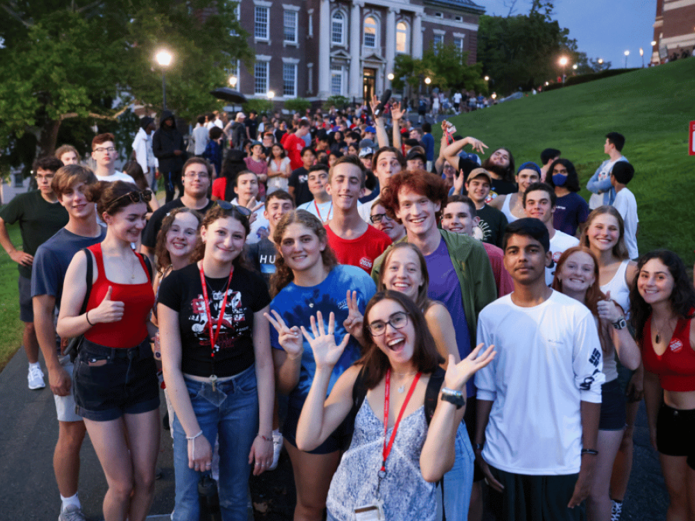 A large group of smiling college students gather on a campus walkway at dusk, posing for a photo while others line up behind them along a grassy hill. A lit academic building with white columns stands in the background as students wave, give thumbs-up, and hold up peace signs.