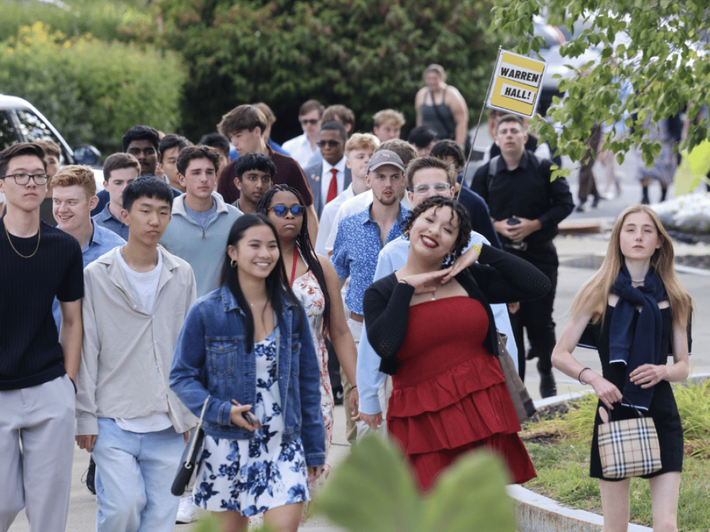 A group of people walking outside on a path amidst greenery. One person holds a sign reading "WARREN HALL." A variety of outfits are visible, reflecting casual styles. The atmosphere appears lively and relaxed.