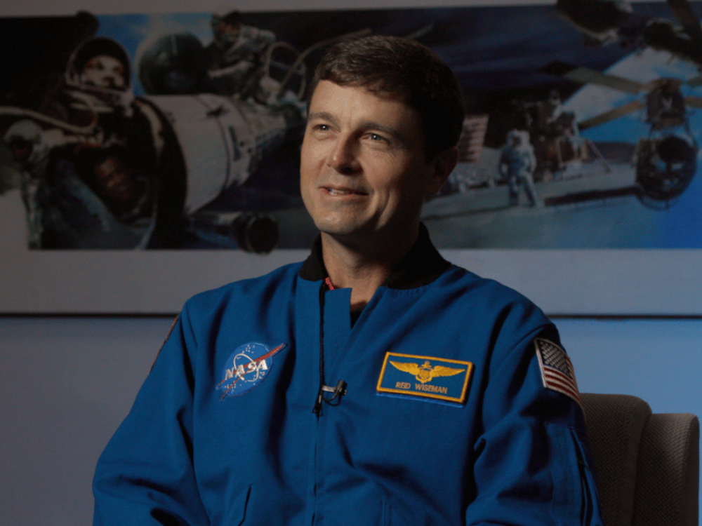 Astronaut in a blue NASA flight suit sits for an interview, smiling slightly. A name patch reading “Reid Wiseman” and a U.S. flag are visible on the suit. Behind him, a wall display shows images of spaceflight, including astronauts, spacecraft, and a space shuttle.